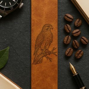 May include: A brown leather bookmark featuring an engraved illustration of a hawk perched on a branch. The bookmark is rectangular and surrounded by coffee beans, a pen, and a watch. The hawk is detailed with intricate lines.