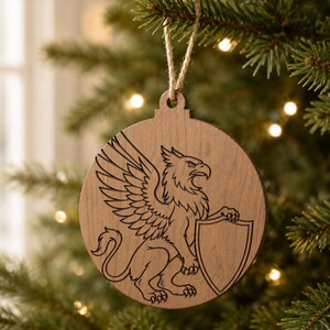 May include: A round, wooden Christmas ornament with a detailed engraving of a griffin holding a shield. The griffin is depicted with a bird's head and wings, and a lion's body. The ornament hangs from a natural twine string.