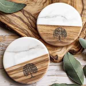 May include: Two round coasters with a half-and-half design. The top half is white marble, and the bottom half is wood. A black etched weeping willow tree design is on the wood. The coasters are on a wooden surface with green leaves.