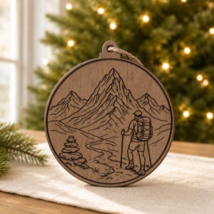 May include: A round wooden ornament with a mountain hiking scene etched into the surface. The design features a hiker with a backpack and walking stick, a mountain range, and a trail. A jute string is attached for hanging.