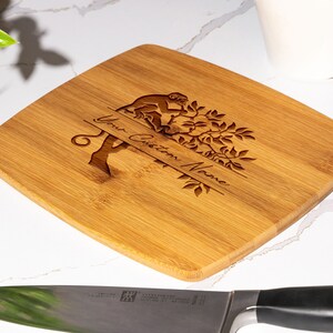 May include: A light brown square-shaped bamboo cutting board with a monkey sitting in a tree design. The board is engraved with the text "Your Custom Name".