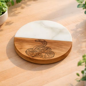 May include: A round serving board with a half-and-half design. The top half is white, and the bottom half is wood-toned. A detailed snake illustration is etched into the wooden portion. The board is surrounded by greenery.
