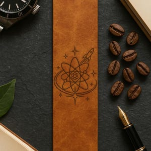 a leather bookmark with a space-themed design, surrounded by coffee beans, a pen, and a watch on a dark surface.