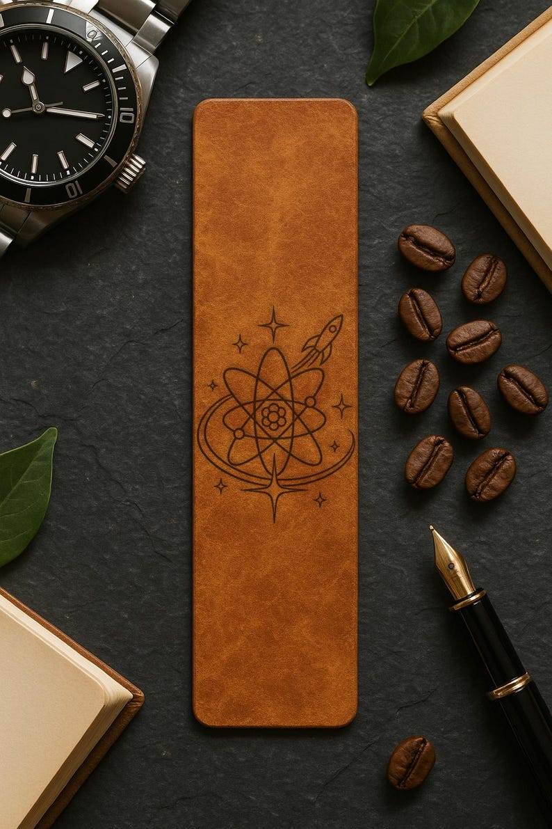 a leather bookmark with a space-themed design, surrounded by coffee beans, a pen, and a watch on a dark surface.