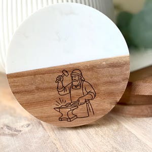May include: A round coaster with a two-tone design. The top half is white, and the bottom half is wood-toned. A black line drawing of a blacksmith at work is etched into the wood. The coaster is likely made of wood and resin.