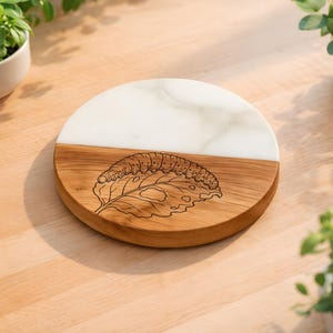 May include: A round wooden and white marble serving board. The wooden section features a detailed engraving of a caterpillar on a leaf. The board is split in half with a white marble top. The board is surrounded by plants.