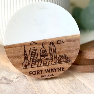 May include: A round coaster with a white top and a wooden bottom. The wooden portion features a laser-etched skyline of Fort Wayne, Indiana, with the city name and state. The coaster is approximately 4 inches in diameter.