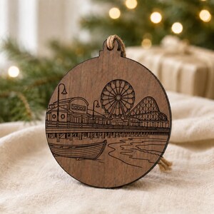A wooden ornament featuring an illustration of a ferris wheel and a boat on a body of water, with a blurred background of greenery and lights.