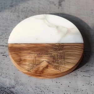 May include: A round coaster with a two-tone design. The top half is white with a marble-like pattern, and the bottom half is wood-grained with an etched image of the Washington Seaside Promenade.