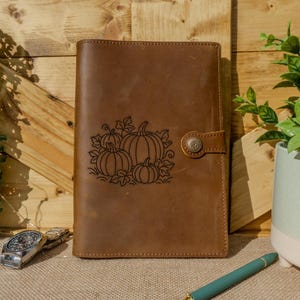 May include: A brown leather journal with a pumpkin design etched on the front. The journal has a button clasp and is closed. A watch and pen are on the surface in front of the journal. The background is a wooden wall.