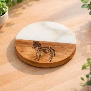 May include: A round serving board with a zebra design. The board is split in half, with a white marble top and a wooden bottom. The zebra is etched into the wood. The board is approximately 8 inches in diameter.