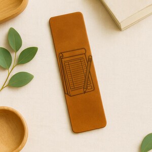 May include: A brown leather bookmark featuring a detailed illustration of a notepad and pencil. The bookmark is rectangular and has rounded edges. The design is in a contrasting dark brown color. The background is a neutral off-white.