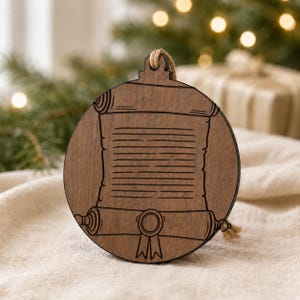 May include: A round, wooden Christmas ornament with a scroll design. The ornament is brown with a detailed engraving of a rolled-up scroll, complete with a ribbon and a loop for hanging. A natural twine loop is attached for hanging.