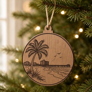 Puede incluir: Adorno navideño redondo de madera con una escena de playa grabada. El diseño incluye una palmera, edificios, un muelle y pájaros. El adorno está colgado de una cuerda de hilo natural.