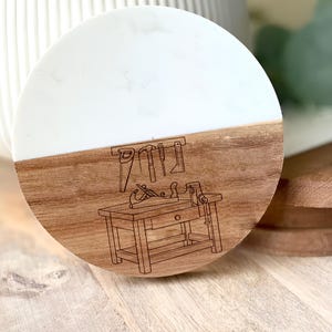 May include: A round coaster with a two-tone design. The top half is white, and the bottom half is wood-toned with a detailed engraving of woodworking tools and a workbench. The coaster is likely made of resin and wood.