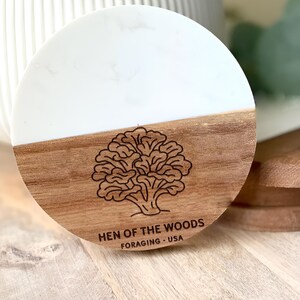 May include: Round wooden coaster with a white marble-like top. The bottom half is wood-toned with a detailed illustration of a mushroom and the text "HEN OF THE WOODS FORAGING - USA".
