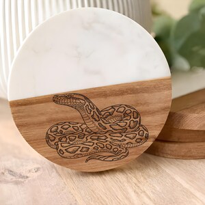 May include: A round coaster with a half-and-half design. The top half is white with a marble pattern, and the bottom half is wood. A detailed engraving of a snake is on the wooden portion. The coaster is approximately 4 inches in diameter.