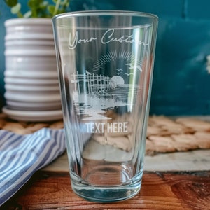 May include: A clear glass pint glass with an etched design of a beach scene with a pier, a sunset, and birds flying. The text "Your Custom Text Here" is etched on the glass.