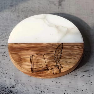 May include: A round coaster with a two-tone design. The top half is white with a marbled effect, and the bottom half is wood-grained. A detailed engraving of an open book, inkwell, and quill pen is on the wooden section. The coaster is a decorative item.