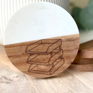 May include: A round coaster with a two-tone design. The top half is white, and the bottom half is wood-toned. A three-tiered tray design is etched into the wood. This coaster is suitable for home or office use.