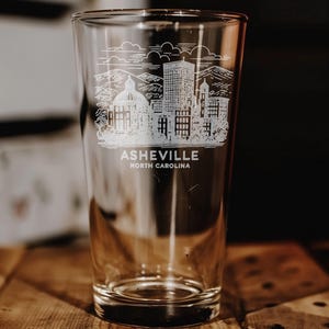 May include: A clear pint glass with a detailed white etched design of the Asheville, North Carolina skyline. The design includes buildings, mountains, and the text "ASHEVILLE NORTH CAROLINA". The glass sits on a wooden surface.