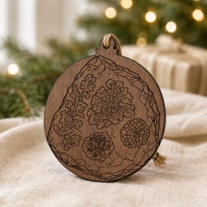 May include: A round, wooden Christmas ornament with a detailed engraved design of various plant-like shapes. The ornament has a natural wood grain finish and a jute string for hanging. The design is a dark brown color.