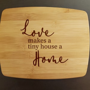 May include: A rectangular bamboo cutting board with rounded corners. The board has a light brown color and features the engraved text "Love makes a tiny house a Home" in a cursive font.