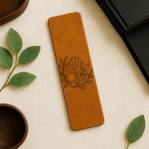 May include: A rectangular, tan leather bookmark with rounded corners. The bookmark features a detailed engraved design of gardening gloves, pruning shears, and leaves. The bookmark is approximately 7 inches long.