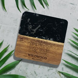 a square coaster with a black marble background and a wooden border, surrounded by green leaves.
