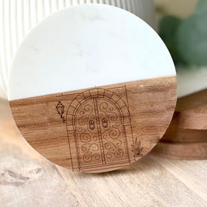 May include: Round coaster with a two-tone design. The top half is white, and the bottom half is wood-toned, featuring an etched illustration of a decorative door. A small lamp and plant are also etched into the wood.