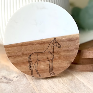 May include: A round coaster with a two-tone design. The top half is white, and the bottom half is wood. A detailed outline of a horse is etched into the wood. The coaster is approximately 4 inches in diameter.