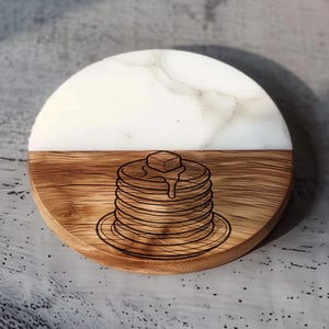 May include: A round coaster with a half-and-half design. The top half is white with a marble effect, and the bottom half is wood-grained. A stack of pancakes with butter is etched into the wood. The coaster is approximately 4 inches in diameter.