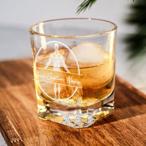 May include: A clear glass tumbler with a white design of a human skeleton with the text "Your Custom Name" on a banner. The glass is filled with a golden liquid and an ice cube.