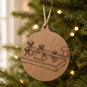 May include: A wooden Christmas ornament with a detailed engraving of a telegraph key. The ornament is round with a hole at the top for hanging. The telegraph key is depicted in black lines against the light brown wood.