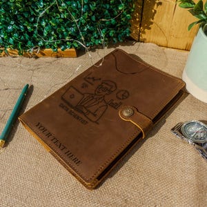 May include: A brown leather journal with a Data Scientist graphic and the text "YOUR TEXT HERE." A silver wristwatch and a teal pen are next to the journal. A small plant in a white and blue pot is in the background.