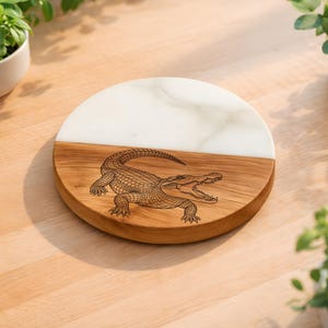 May include: A round serving board with a half-and-half design. The top half is white, and the bottom half is wood-toned. An alligator illustration is etched into the wood. The board is surrounded by plants in white pots.