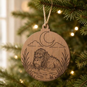 May include: Wooden Christmas ornament with a laser-etched design. The ornament features a creature in water, a crescent moon, clouds, and reeds. The ornament hangs from a natural twine string.