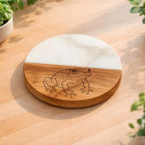 May include: A round wooden and white marble serving board with a detailed etched illustration of a toad. The board is split in half, with the top half in white marble and the bottom half in wood.