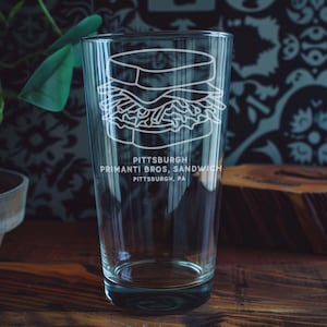 May include: A clear glass with a sandwich design etched on the front. The text "PITTSBURGH PRIMANTI BROS, SANDWICH PITTSBURGH, PA" is below the sandwich illustration. The glass is on a wooden surface.
