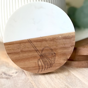 May include: A round wooden coaster with a white top half and a brown bottom half. The bottom half has a detailed engraving of a gardening fork and basket. The coaster is next to a wooden serving board.