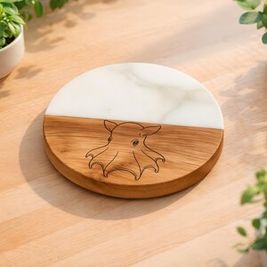 May include: A round wooden and white marble serving board. The board is divided in half, with a light wood grain on the bottom half and white marble on the top half. An octopus design is etched into the wood. The board is on a light wood surface.