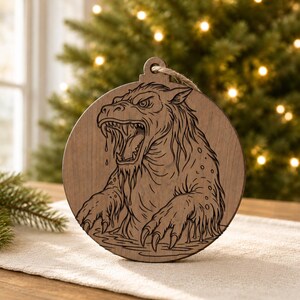 May include: Wooden Christmas ornament featuring a detailed engraving of a ferocious, mythical creature with sharp teeth and claws. The round ornament has a natural wood grain finish and a jute string for hanging. The creature is depicted in a water-like setting.