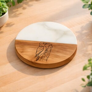 May include: A circular serving board featuring a split design with one half made of white marble and the other half made of natural brown wood. The wooden section is engraved with a detailed black line drawing of a bird perched on a tree trunk.