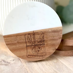 May include: A round coaster with a two-tone design. The top half is white, and the bottom half is wood-toned. A line drawing of stationery, including a pencil, pen, and ruler, is etched into the wood. The coaster is approximately 4 inches in diameter.