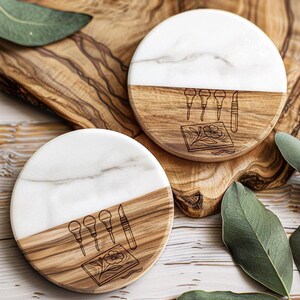 May include: Two round coasters. Each coaster has a white marble top and a wood-grain bottom. The wood-grain section has a carved design of tools and a floral pattern. The coasters are on a wooden cutting board.