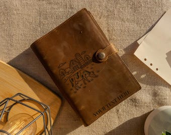 May include: A brown leather journal with a farm scene illustration and the text "YOUR TEXT HERE". The journal has a button closure and is lying on a textured surface. A wooden cutting board and a light bulb are in the background.