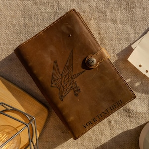 May include: A brown leather journal with a geometric eagle design and the text "YOUR TEXT HERE". The journal has a button closure and is lying on a textured surface. A light bulb in a metal cage is in the lower left corner.