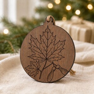 May include: A round, wooden ornament with a maple leaf design. The ornament is brown with a natural wood grain and a jute string for hanging. The leaf design is etched into the wood.