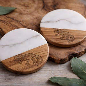 May include: Two round coasters with a white marble top and a wood base. Each coaster features a laser-etched gecko design. The coasters are set on a wooden surface with green leaves.