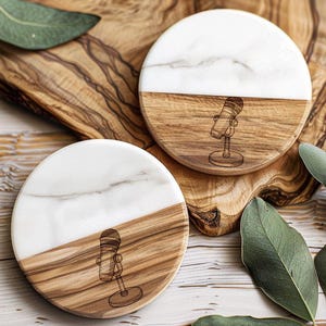 May include: Two round coasters with a microphone design. Each coaster is split diagonally, with a white marble top and a wooden bottom. The microphone is etched into the wood. The coasters are on a wooden surface.